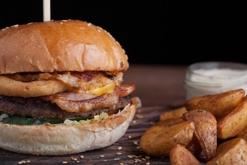 Closeup of a tasty Burger with snacks in the form of potatoes with white garlic sauce. Juicy Burger with onion rings, bacon and cheese on a dark wood background