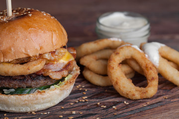 Closeup of a tasty Burger with appetizers such as fried onion rings with a white garlic sauce. Juicy Burger with bacon and cheese on a dark wood background