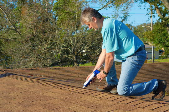 Homeowner Patching Roof With Roof Repair Tar In Caulk Gun Doing Home Maintenance To Protect The House From Rain, Wind, Storms And Hurricanes.