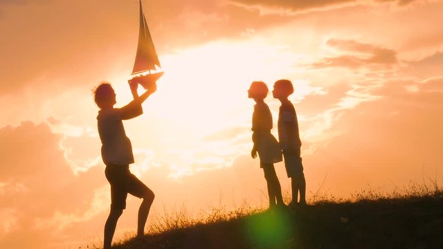 Model of a sailboat. A man is holding a ship model against the sky. The father gives the ship to the child. Backlight. Silhouettes of people against the sky and the sun.
