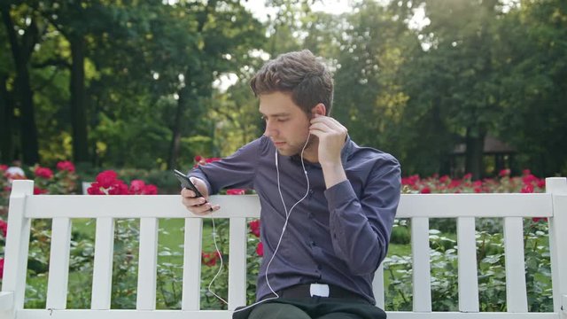 A young man sitting on a white bench in the park using a phone listening to music. Medium shot