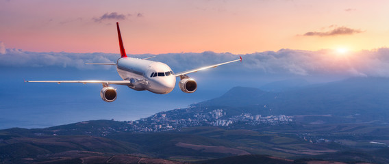Passagiers vliegtuig. Landschap met wit vliegtuig vliegt in de oranje lucht met wolken over bergen, zee bij kleurrijke zonsondergang. Passagiersvliegtuigen landen. Commercieel vliegtuig. Prive jet. Reis