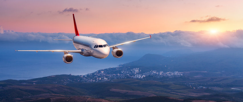 Passenger airplane. Landscape with white airplane is flying in the orange sky with clouds over mountains, sea at colorful sunset. Passenger aircraft is landing. Commercial plane. Private jet. Travel © den-belitsky