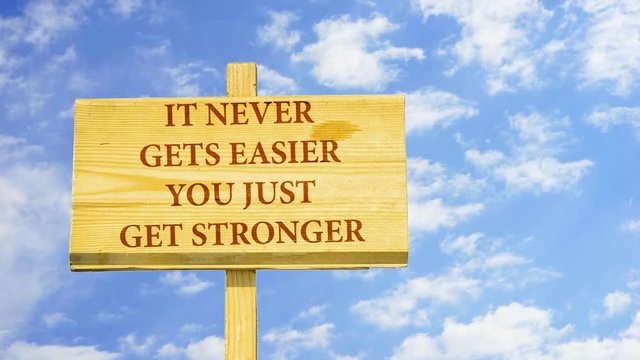 It never gets easier. You just get stronger. Words on a wooden sign against time lapse clouds in the blue sky.