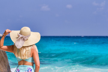 A girl in a blue summer dress with a hat on his head, standing on white sand and looking at the ocean