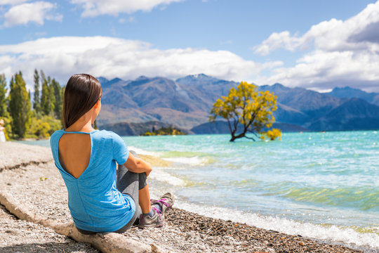 New Zealand travel tourist woman looking at Wanaka lone tree lake landscape. Girl relaxing enjoying view sitting on beach at Wanaka nature mountains outdoors.
