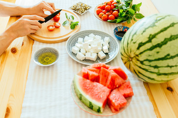 Close up of female housewife hands cutting cherry tomatoes on wooden cutting board on wooden table with watermelon, pieces, chees feta, pumpkin seeds, mint, tomatoes, side view