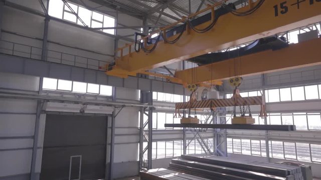 Yellow overhead crane with electromagnetic beam grippers carries steel cargo in engineering plant shop warehouse.