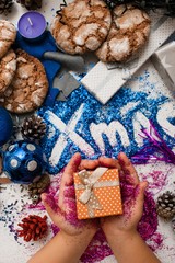 Christmas festive background of gifts top view. Little kid holds small present in purple glittering hands on bright xmas inscription and handmade decoration backdrop. Holiday and celebration concept