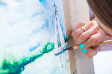 Young dark-haired woman painter watercolor painting and wooden brush on white paper seascape with a single-deck yacht