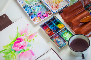 Drawing album with painted peonies and dog roses, leather case with brushes, watercolor palette, pencils and a mug with water on a white table, top view