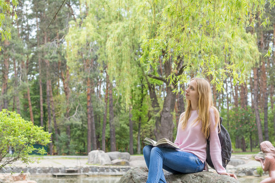 Cute romantic blond teenage-girl is reading a book sitting under the tree in the city park in the sunny weather. A pond surrounded by rocks with trees reflexion in the water is at the background.