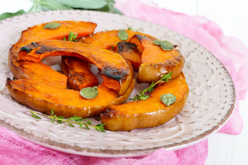 Sliced of baked pumpkin in a honey sauce with aromatic herbs on a ceramic plate on a white wooden background.