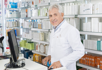 Senior Chemist Using Computer At Counter In Pharmacy