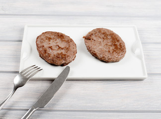 Burgers on white plate next to knife and fork on wooden table. Food.