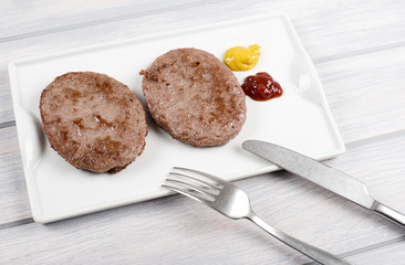 Burgers on white plate next to knife and fork with ketchup and mustard on wooden table. Food.