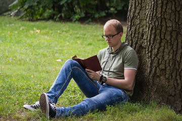 Young man in glasses reading and writing in notebook sitting on the grass near the tree in park. Maybe student or writer