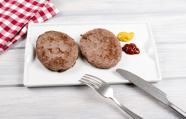 Burgers on white plate next to napkin, knife and fork with ketchup and mustard on wooden table. Food.