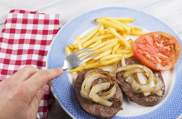 A hand with fork on french fries and hamburger with onion and tomato on blue plate next to napkin on wooden table. Food.