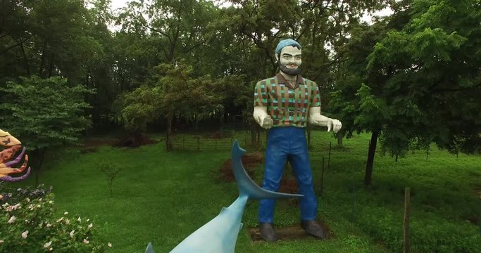 Camera orbiting above Paul Bunyan the muffler man and Finding Nemo shark all set in the countryside.