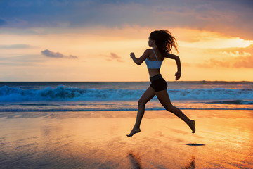 Fototapeta na wymiar Barefoot sporty girl silhouette running along ocean surf by water pool to keep fit and health. Sunset black beach background with sun. Woman fitness, jogging sport activity on summer family holiday.