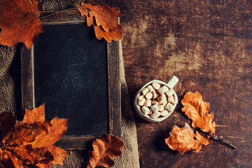 Autumn, fall leaves on vintage Empty blank black chalkboard with hot cup of coffee, marshmallows  and  copyspace. Cozy Autumn Still Life Background. Autumn red and orange leaves