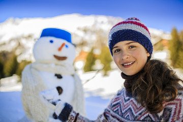 Happy smiling teenage girl playing with a snowman on a snowy winter village in mountains, Switzerland