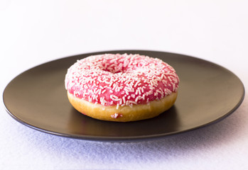 Brightly pink donut on a black ceramic plate on white background.