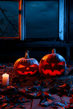 The concept of Halloween. Two luminous evil scary pumpkins, jack-lantern, with candles, leaves and eerie branches outside the window with a warm and cold blue light on the wooden floor