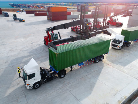 aerial view of loading forklift stacker handling cargo container lifting to trailers in import export logistic zone, with international global port in background