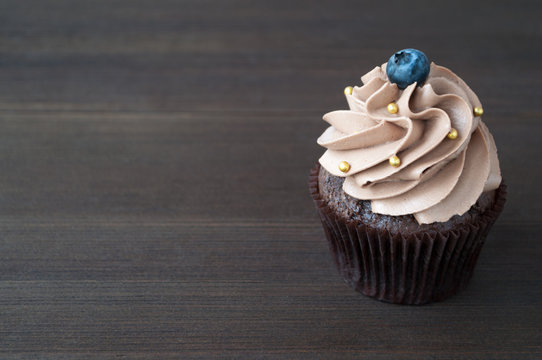 Chocolate cupcake with whipped chocolate cream, decorated fresh blueberry, gold confectionery sprinkling on dark wooden table. Picture for a menu or a confectionery catalog with copy space.