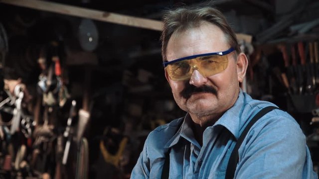 An adult carpenter wearing a uniform and protective goggles in a small carpentry workshop, against a wall with tools.