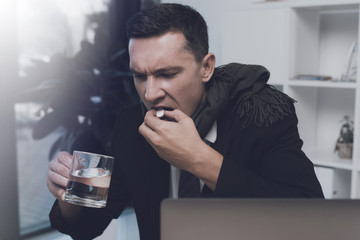 A sick man is sitting at his workplace in the office. He has a pill and a cup of water in his hands