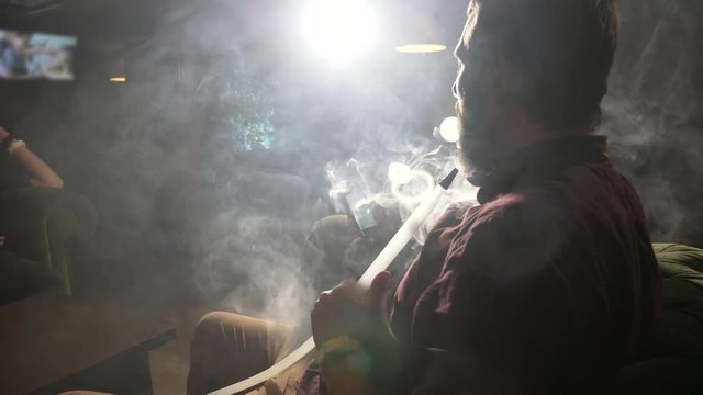 Young men using smart phone and smoking hookah at dark shisha lounge