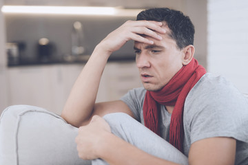 A man with a cold sits on the couch, hiding behind a red rug. He has a headache