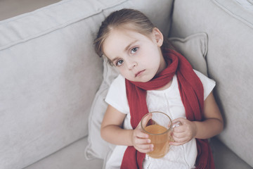 Little sick girl sits on a white couch wrapped in a red scarf. She is sitting with a cup of medicinal tea
