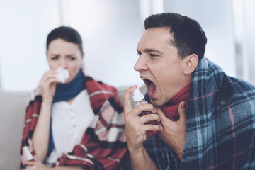 The couple is sitting on the couch wrapped in blankets. The man uses a spray for the throat