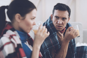 The couple is sitting on the couch wrapped in blankets. Man and woman are sick. They drink medicinal tea