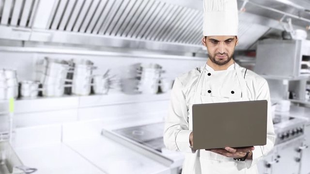Young Caucasian chef using a laptop computer while wearing uniform and standing in the kitchen