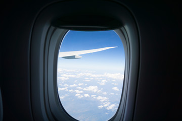 Outlook through plane window while traveling with window surround framing blue sky and clouds with focus on wing outside.