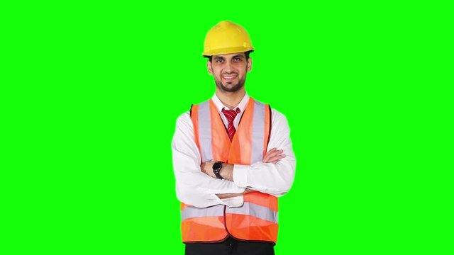 Confident Arabian engineer standing in the studio while crossed hands and wearing safety vest with helmet in front of green screen background