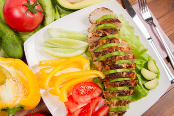 Chicken fillet with avocado and some vegetable on the side (tomato, cucumber, bell pepper) on a wooden background