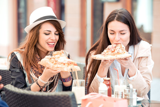 Two cheerful girls eating pizza in a outdoor cafe.