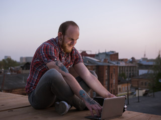Fototapeta na wymiar Pleasant social media communication on roof. Happy smiling male freelancer with laptop outdoors in focus on foreground, creative self-education, urban background with free space