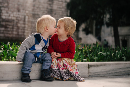 Two small children sitting on a low wall share a kiss.