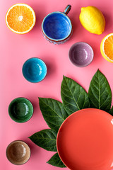 Colored cups and plates pattern decorated by exotic leaves and fruits on pink background top view