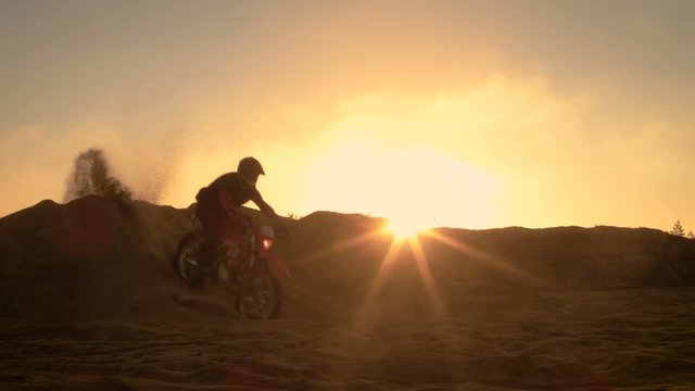 Professional Motocross Motorcycle Rider Drives Over the Dune and Further Down the Off-Road Track. It's Sunset and Track is Covered with Smoke/ Mist. Shot on RED EPIC-W 8K Helium Cinema Camera.