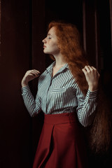 Sensual young red haired model wearing white striped shirt and red skirt. Woman posing in a dark room