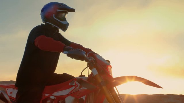 Professional FMX Motorcycle Rider Prepares to Start Driving on His Bike Over Hard Sandy Off-Road Terrain. Sun is Setting. Shot on RED EPIC-W 8K Helium Cinema Camera.