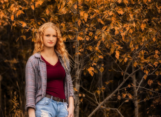 The upper body of a teenage girl in a tank top and blouse and ripped jeans in front of autumn colored trees